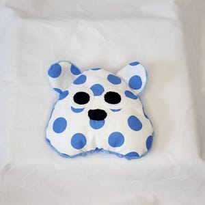 May include: A handmade, white plush toy shaped like a bear's head, adorned with large blue polka dots. The toy has black embroidered eyes and nose, with blue trim around the edges and ears. The background is a white surface.