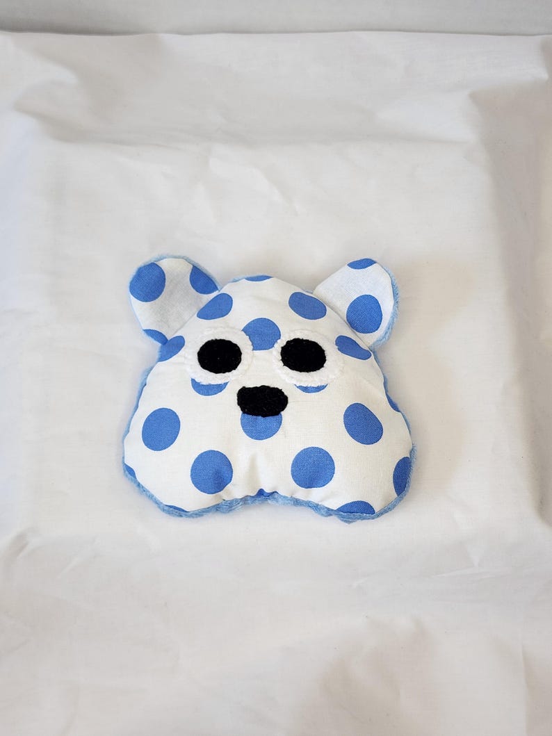 May include: A handmade, white plush toy shaped like a bear's head, adorned with large blue polka dots. The toy has black embroidered eyes and nose, with blue trim around the edges and ears. The background is a white surface.