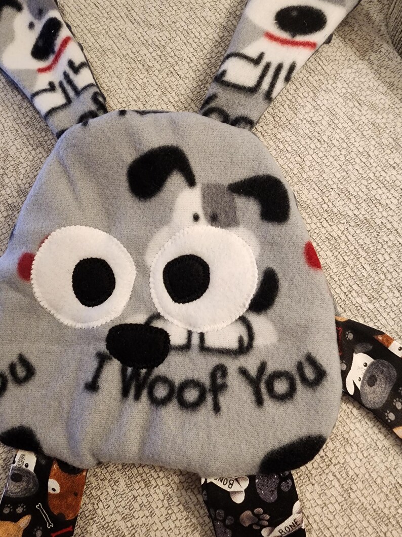 May include: A gray plush dog toy with the text "I Woof You". The toy has large white eyes with black pupils, black ears, and red accents. The toy has multiple patterned fabric appendages.