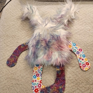 May include: A handmade plush toy with a fluffy, multi-coloured body in shades of purple, blue, and pink. The arms and legs are made of patterned fabric with floral and swirl designs. The toy is designed to be soft and cuddly.