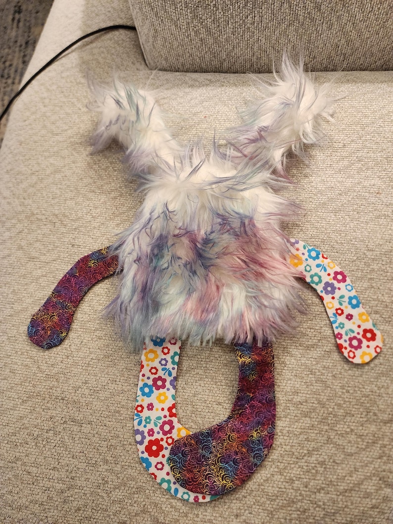May include: A handmade plush toy with a fluffy, multi-coloured body in shades of purple, blue, and pink. The arms and legs are made of patterned fabric with floral and swirl designs. The toy is designed to be soft and cuddly.