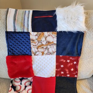 May include: A patchwork pillow featuring various textures and patterns. The design includes baseballs, stars, and solid colors like red, blue, and white. One section has a zipper. The pillow is made of soft fabrics.