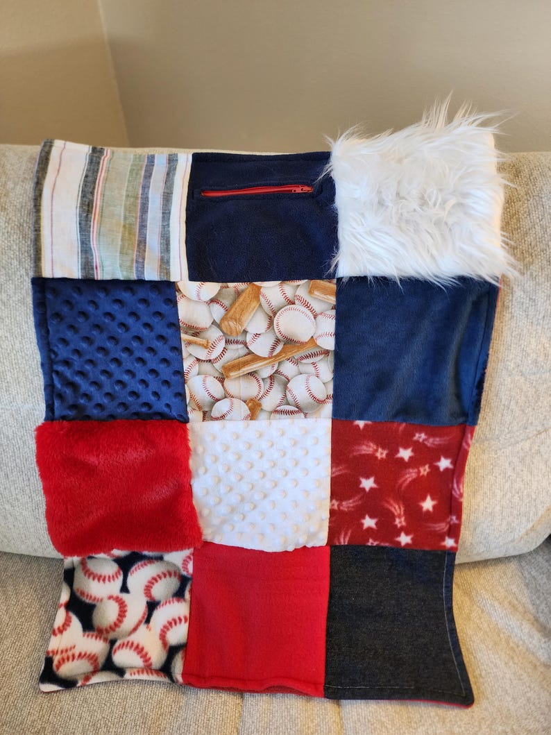 May include: A patchwork pillow featuring various textures and patterns. The design includes baseballs, stars, and solid colors like red, blue, and white. One section has a zipper. The pillow is made of soft fabrics.