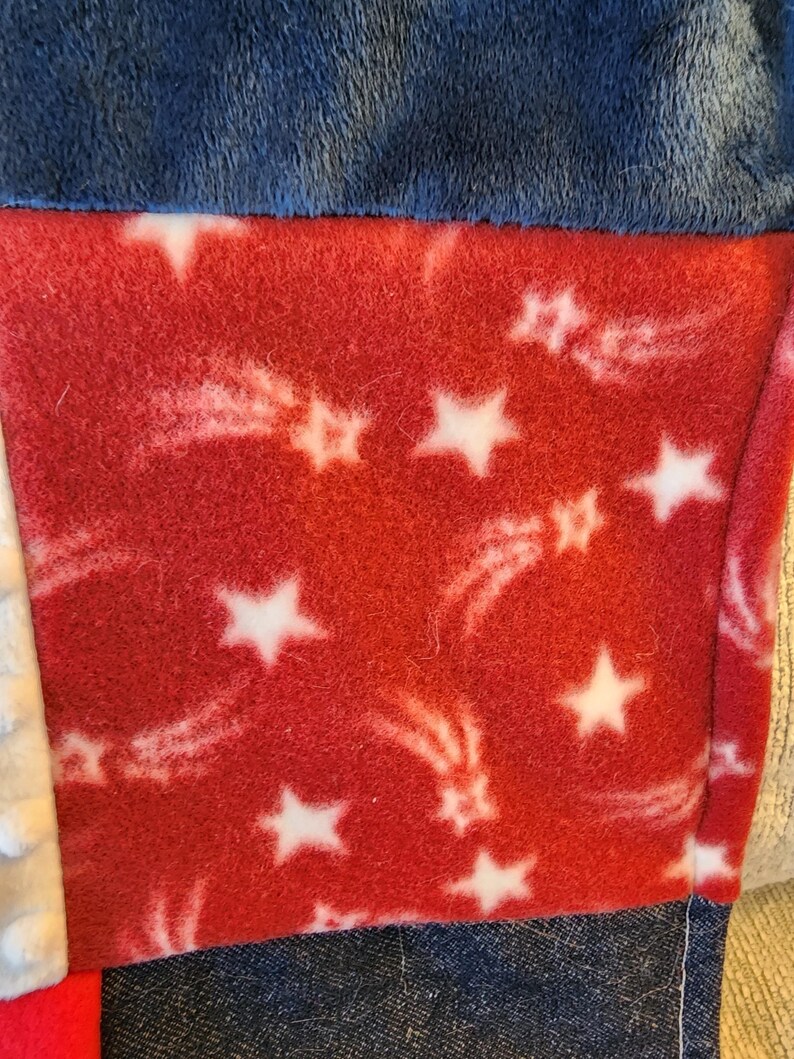 May include: Close-up of a patchwork textile featuring a red fleece panel with white stars and shooting star designs. The panel is bordered by blue and denim fabrics, creating a patriotic theme.