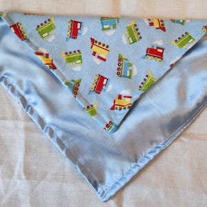 May include: A baby blanket with a light blue satin border and a patterned center. The center fabric features colorful train illustrations on a light blue background. The blanket is folded in a triangle shape.