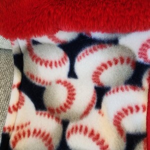 May include: A baseball-themed fleece blanket with a red plush trim. The blanket features a pattern of white baseballs with red stitching on a navy blue background. The top edge is a vibrant red.