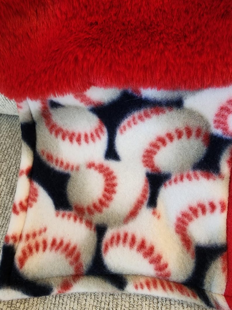 May include: A baseball-themed fleece blanket with a red plush trim. The blanket features a pattern of white baseballs with red stitching on a navy blue background. The top edge is a vibrant red.