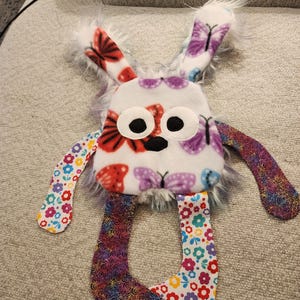 May include: A handmade plush bunny toy with a white body decorated with red and purple butterfly patterns. The bunny features large black eyes, fluffy white ears, and colourful floral and rainbow patterned limbs. A children's toy.