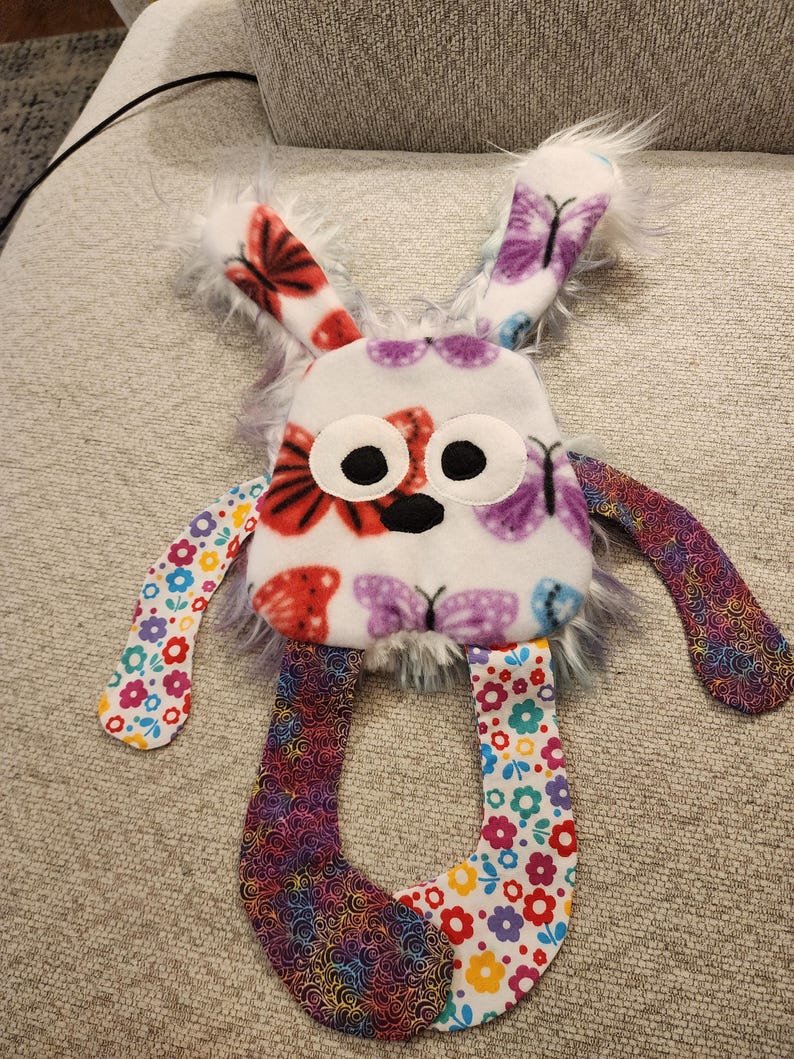 May include: A handmade plush bunny toy with a white body decorated with red and purple butterfly patterns. The bunny features large black eyes, fluffy white ears, and colourful floral and rainbow patterned limbs. A children's toy.
