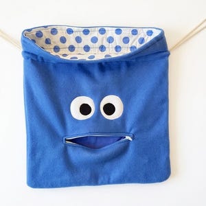 May include: A blue fabric drawstring bag featuring a character face with two large white eyes with black pupils and a horizontal zippered mouth. The interior lining of the bag is white with a blue polka dot pattern and beige drawstrings.