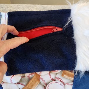 May include: A navy blue fabric with a red zipper pocket, baseball-themed fabric, and white faux fur trim. The baseball fabric features white baseballs and brown baseball bats. The pocket is being opened by a hand.