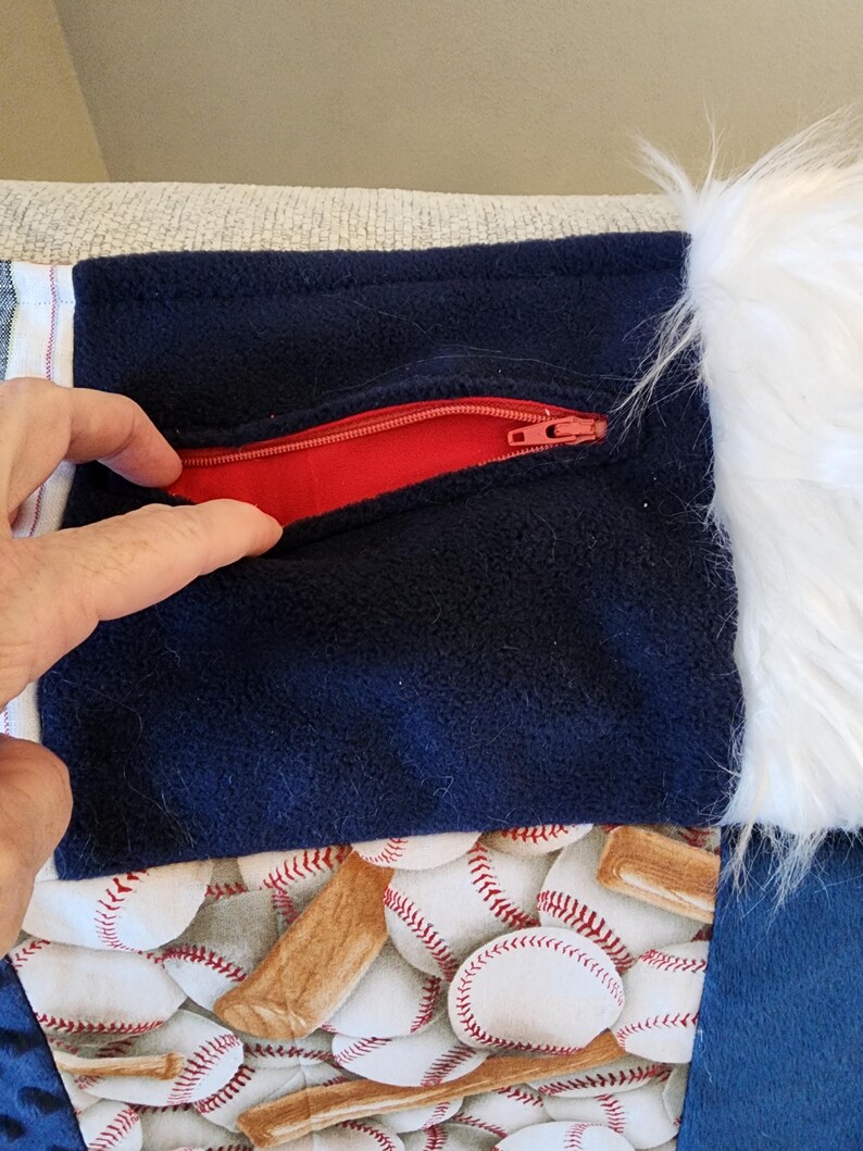 May include: A navy blue fabric with a red zipper pocket, baseball-themed fabric, and white faux fur trim. The baseball fabric features white baseballs and brown baseball bats. The pocket is being opened by a hand.