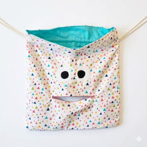 May include: A drawstring bag featuring a white fabric with a colorful pattern of small triangles in pink, yellow, and blue. The bag has two large white circular eyes with black pupils and a horizontal zippered mouth opening. The interior is teal.