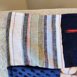 May include: A handmade patchwork blanket featuring various fabrics. The top section has a striped pattern in white, black, red, and green. Below is a navy blue, dimpled fabric, and a section with baseballs. A red zipper is visible.