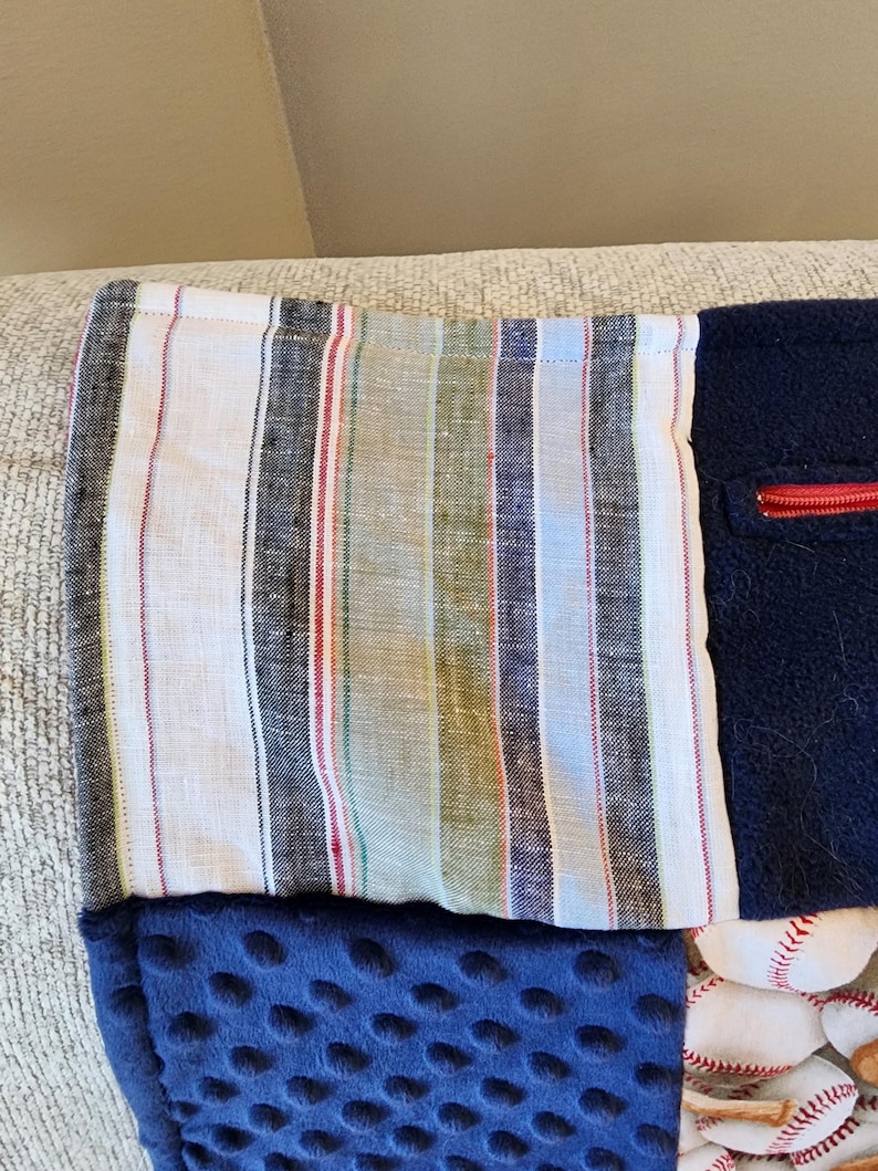 May include: A handmade patchwork blanket featuring various fabrics. The top section has a striped pattern in white, black, red, and green. Below is a navy blue, dimpled fabric, and a section with baseballs. A red zipper is visible.
