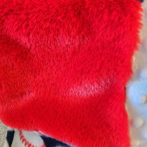 May include: A close-up of a red, plush, rectangular item, possibly a blanket or pillow, with a textured surface. The red item is layered over a navy blue fabric with raised dots and a baseball-themed fabric.