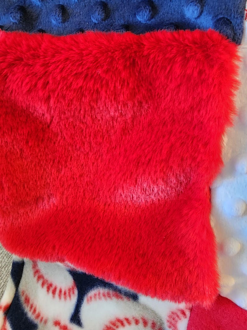 May include: A close-up of a red, plush, rectangular item, possibly a blanket or pillow, with a textured surface. The red item is layered over a navy blue fabric with raised dots and a baseball-themed fabric.