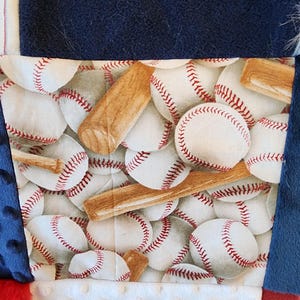 May include: A patchwork quilt featuring a baseball theme. The quilt has a central panel with baseballs and bats, surrounded by navy blue, red, and white fabrics. The baseballs are white with red stitching.