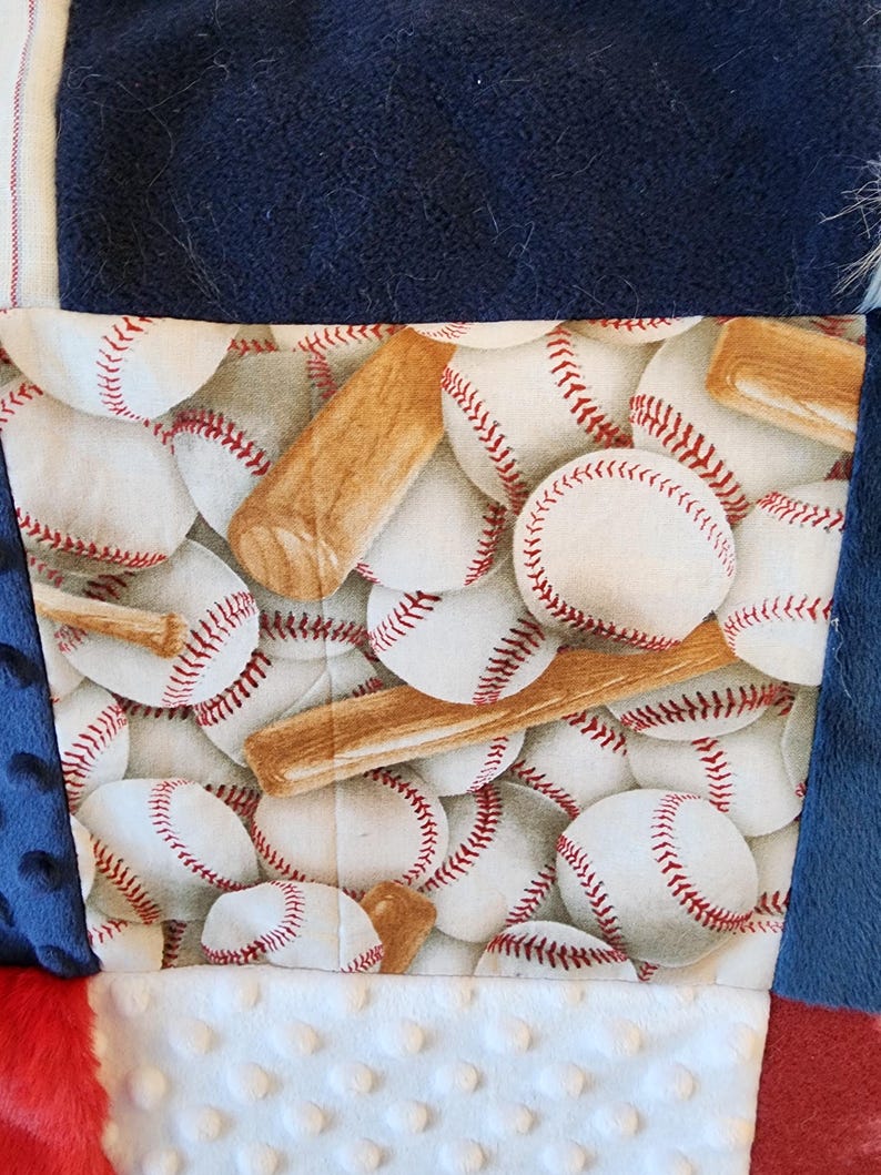 May include: A patchwork quilt featuring a baseball theme. The quilt has a central panel with baseballs and bats, surrounded by navy blue, red, and white fabrics. The baseballs are white with red stitching.