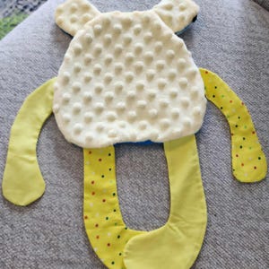 Op de afbeelding: Een zacht babyspeelgoed in de vorm van een vriendelijk wezen. Het lichaam is cr&egrave;mekleurig met een getextureerd oppervlak, en de armen en benen zijn geel met kleurrijke stippen. Het speelgoed heeft kleine, afgeronde oren.