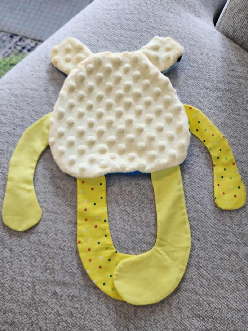 Op de afbeelding: Een zacht babyspeelgoed in de vorm van een vriendelijk wezen. Het lichaam is cr&egrave;mekleurig met een getextureerd oppervlak, en de armen en benen zijn geel met kleurrijke stippen. Het speelgoed heeft kleine, afgeronde oren.