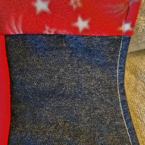 May include: Close-up of a section of a sewn item. The top portion is red fleece with white stars. Below is a section of dark blue denim fabric. The two fabrics are sewn together with white thread.