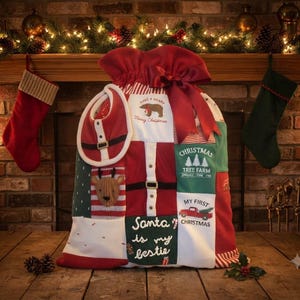 May include: A large fabric Christmas gift sack featuring a patchwork design of red, white, and green panels. The sack includes a red ribbon bow, a baby bib, and printed text such as "Have a Beary Merry Christmas," "Christmas Tree Farm," "My First Christmas," and "Santa is my bestie."