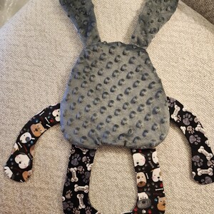 May include: A gray plush bunny-shaped baby toy with textured ears and body. The arms and bottom loop feature a black fabric with a pattern of dog faces, paw prints, and bones. The toy is designed for babies.