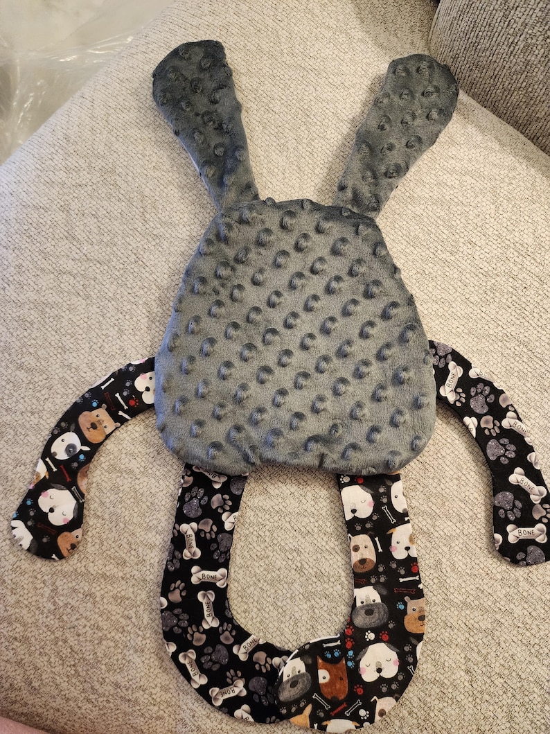 May include: A gray plush bunny-shaped baby toy with textured ears and body. The arms and bottom loop feature a black fabric with a pattern of dog faces, paw prints, and bones. The toy is designed for babies.