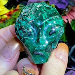 May include: A green malachite alien head sculpture with detailed facial features. The sculpture is held in a hand with rings. The background features flowers in shades of yellow, purple, and black.