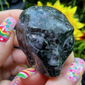 Op de afbeelding: Een donkergrijze alienhoofdsculptuur met gedetailleerde gelaatstrekken. De sculptuur wordt vastgehouden in een hand met kleurrijke nail art en diverse ringen. De achtergrond toont groen gebladerte en een gele bloem.