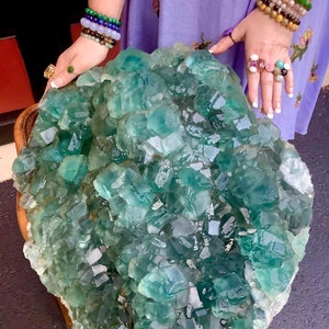 May include: A large, green fluorite crystal cluster with many individual crystals. The crystal cluster is sitting on a wooden table.