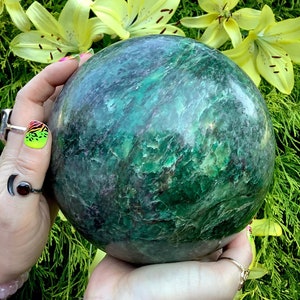 Könnte beinhalten: Eine große, polierte Kugel aus Aventurinkristall in Grün. Die Kugel hat ein wirbelndes, marmoriertes Muster und wird in den Händen einer Person gehalten.