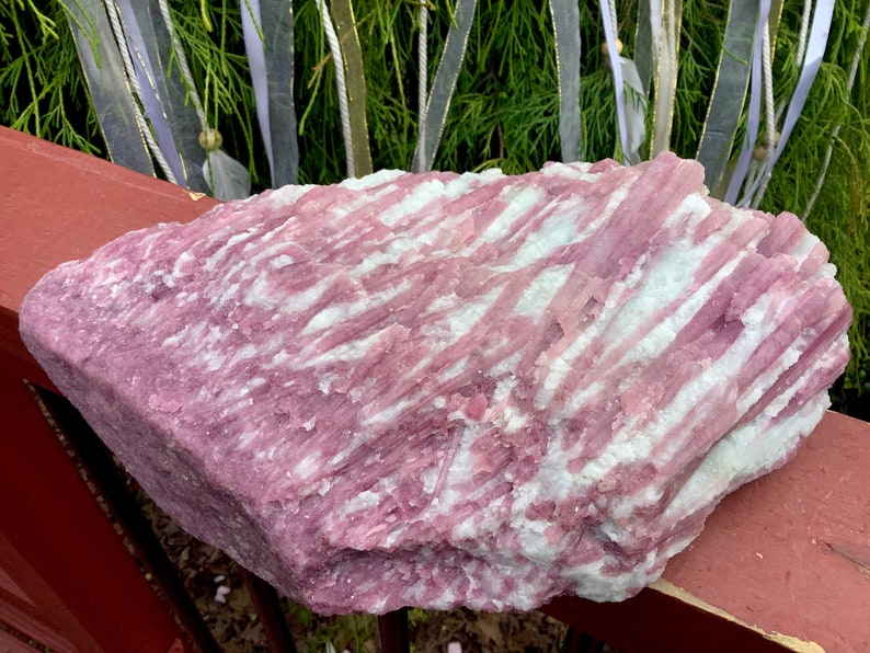 May include: A large, raw mineral specimen featuring a striking combination of vibrant pink and opaque white crystalline structures. The stone displays a layered, fibrous texture with elongated, parallel formations running across its surface.