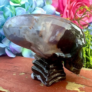 May include: A polished skull sculpture in shades of brown, white, and gray, resting on a dark, layered base. The skull's surface reflects light, showcasing its intricate details. The sculpture is set against a backdrop of colorful flowers.