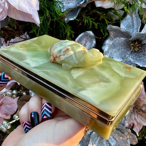 Pode incluir: Uma caixa retangular de ônix verde claro com acabamento dourado e uma figura de tartaruga esculpida no topo. A caixa tem uma aparência marmorizada e está colocada em um fundo de flores e vegetação.