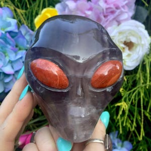 May include: A dark purple alien head sculpture with red eyes. The sculpture is held in a hand with turquoise nail polish. The background features colorful flowers and greenery. The alien head is a unique decorative item.