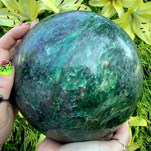 Puede incluir: Una gran piedra esférica pulida que presenta una paleta de colores verde intenso con texturas veteadas y sutiles inclusiones de vetas minerales de color púrpura oscuro y negro en toda la superficie, mostrando una forma natural lisa.