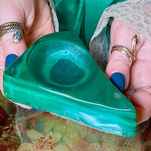 May include: A triangular-shaped bowl carved from polished green malachite stone, featuring distinct dark and light green banded patterns throughout the surface. The bowl has a smooth, concave center and a thick, solid base with natural stone textures.