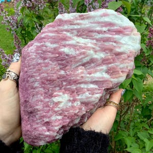 May include: A large, raw mineral specimen featuring a textured surface with alternating bands of vibrant pink and opaque white crystalline structures. The stone displays a jagged, natural formation with visible striations throughout the rock.