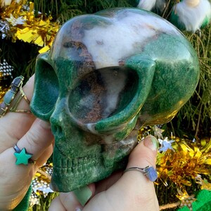 May include: A green and brown gemstone skull sculpture. The skull is held in hands, showcasing its polished surface and detailed features. The sculpture has a natural, earthy aesthetic.
