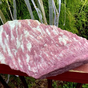 May include: A large, raw mineral specimen featuring a striking combination of vibrant pink and opaque white crystalline structures. The stone displays a rough, textured surface with elongated, parallel formations throughout the entire piece.