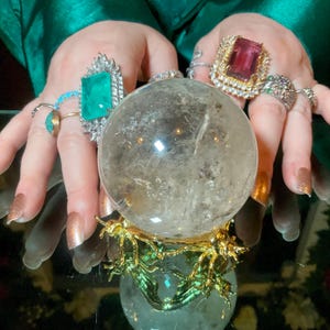 Puede incluir: Una bola de cristal transparente sobre un soporte decorativo dorado y verde. Las manos que sostienen la bola están adornadas con varios anillos, incluido un gran anillo con corte esmeralda y un anillo con una piedra preciosa rosa. Las uñas están pintadas de dorado.