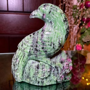 May include: A carved stone figurine of a bird perched on a base with two floral shapes. The sculpture is primarily green with prominent black mineral inclusions and small patches of vibrant magenta throughout the polished stone surface.