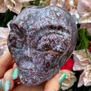 May include: A carved stone sculpture of a human-like face. The sculpture is primarily burgundy with blue and white veining. The face has closed eyes and a serene expression. The sculpture is held in a person's hand.