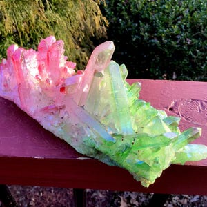 May include: A cluster of raw quartz crystals featuring a vibrant gradient colour transition from bright pink on the left side to a luminous lime green on the right side. The mineral formation has multiple sharp, pointed, and translucent crystal points.