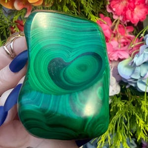 May include: A polished malachite stone featuring vibrant green hues and distinct, swirling concentric banding patterns. The mineral has a smooth, rounded surface with a glossy finish that highlights the natural, intricate layers of the stone.