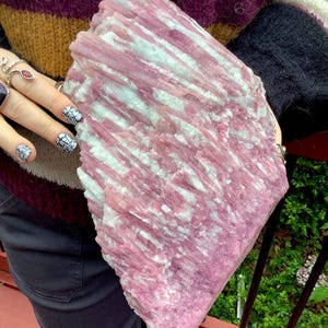 May include: A large, raw mineral specimen featuring a vertical, striated crystalline structure. The stone displays a prominent color palette of vibrant magenta and soft pink hues, interspersed with streaks of opaque white mineral deposits.