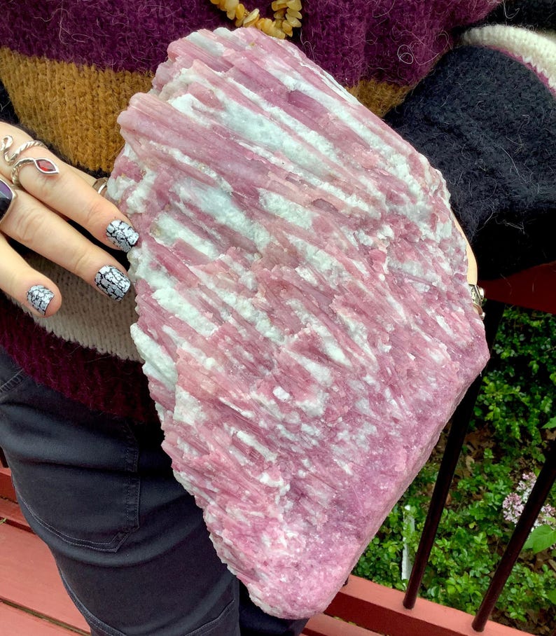 May include: A large, raw mineral specimen featuring a vertical, striated crystalline structure. The stone displays a prominent color palette of vibrant magenta and soft pink hues, interspersed with streaks of opaque white mineral deposits.