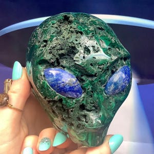 May include: A green malachite alien head sculpture with blue lapis lazuli eyes. The sculpture has a rough, textured surface and is held in a hand with turquoise nails. The background is a blurred blue and white.